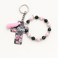 Light pearl pink and black beaded stretch bracelet with silver cross charm beside a dark cross tag keychain with pink florals, Be Strong lettering, and pale pink tassel