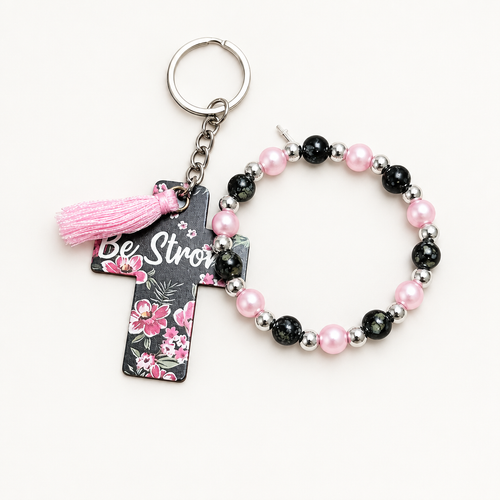 Light pearl pink and black beaded stretch bracelet with silver cross charm beside a dark cross tag keychain with pink florals, Be Strong lettering, and pale pink tassel