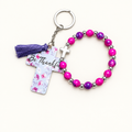 Magenta and purple beaded stretch bracelet with silver cross charm beside a white cross tag keychain with pink florals, Be Thankful lettering, and purple tassel