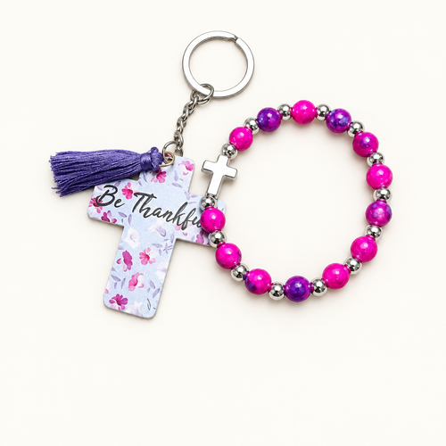 Magenta and purple beaded stretch bracelet with silver cross charm beside a white cross tag keychain with pink florals, Be Thankful lettering, and purple tassel