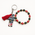 Red, black, and gold beaded stretch bracelet with silver cross charm beside a black cross tag keychain with red florals, Believe lettering, and red tassel