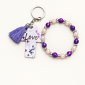Purple and pearl beaded stretch bracelet with silver cross charm beside a white cross tag keychain with purple florals, Love lettering, and purple tassel