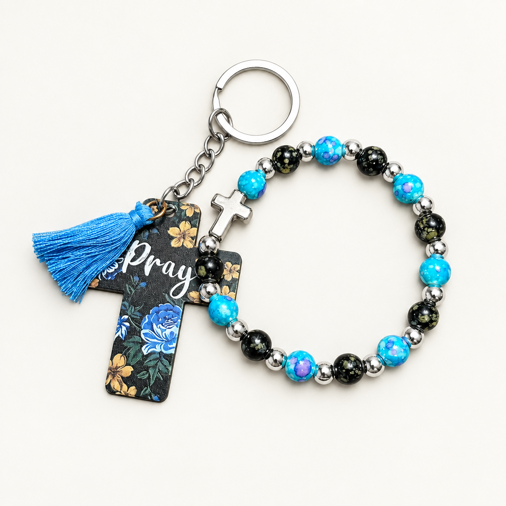Teal and black beaded stretch bracelet with silver cross charm beside a black cross tag keychain with teal florals, Pray lettering, and teal tassel